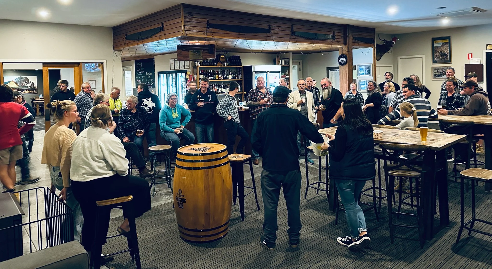 People gathered in a bar or brewery enjoying drinks and socializing, with some standing and some sitting, in a cozy setting with wooden decor.