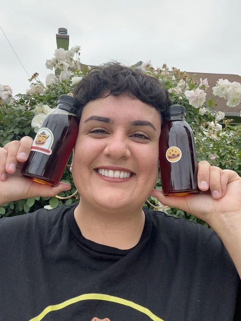 Person smiling and holding two jars with emoji stickers in front of flowering bushes outside.
