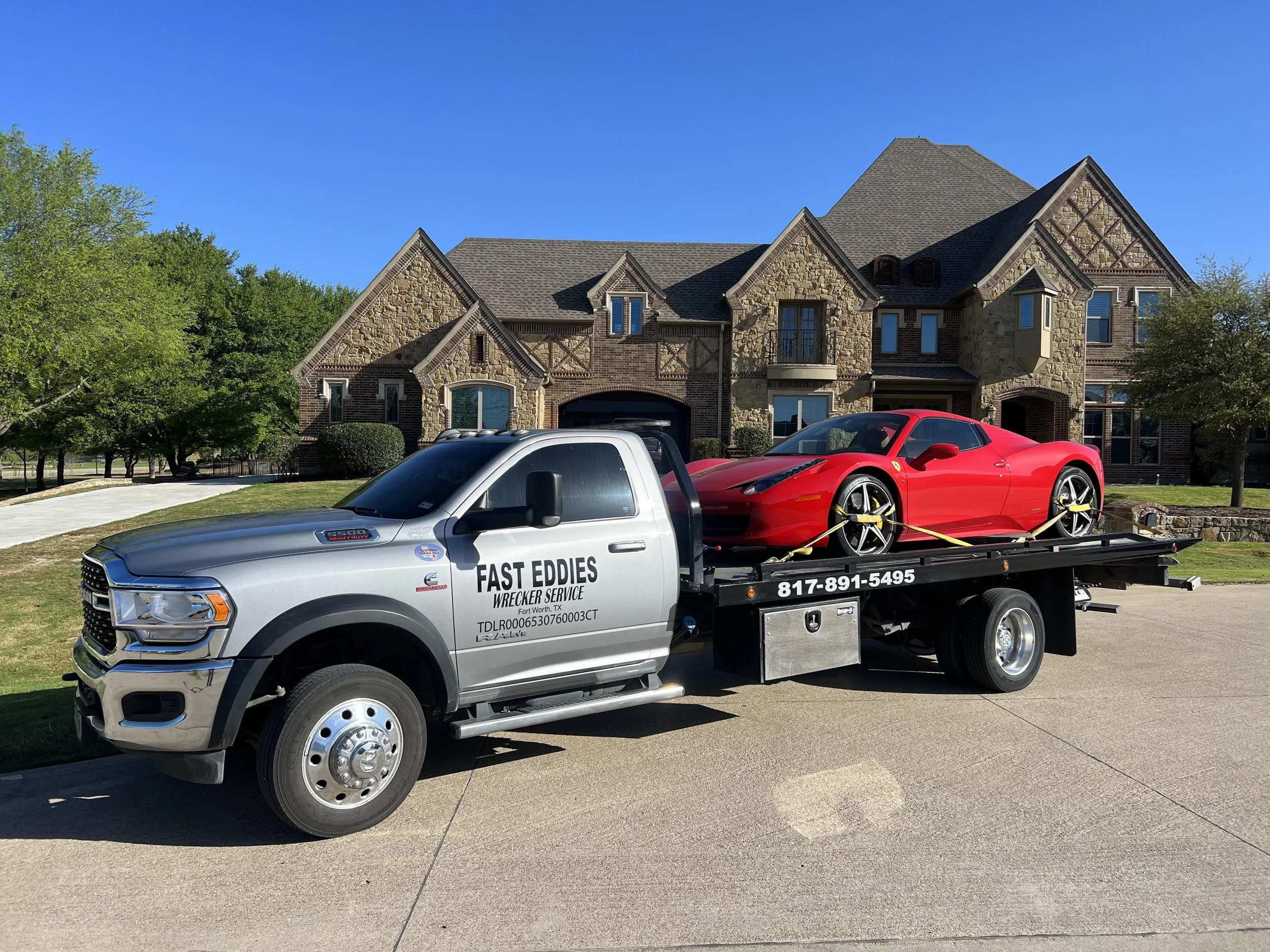 A silver tow truck with black lettering reading 'Fast Eddies Wrecker Service' on the side, carrying a red sports car, in front of a large, multi-story house with stone and brick exterior under a clear blue sky.
