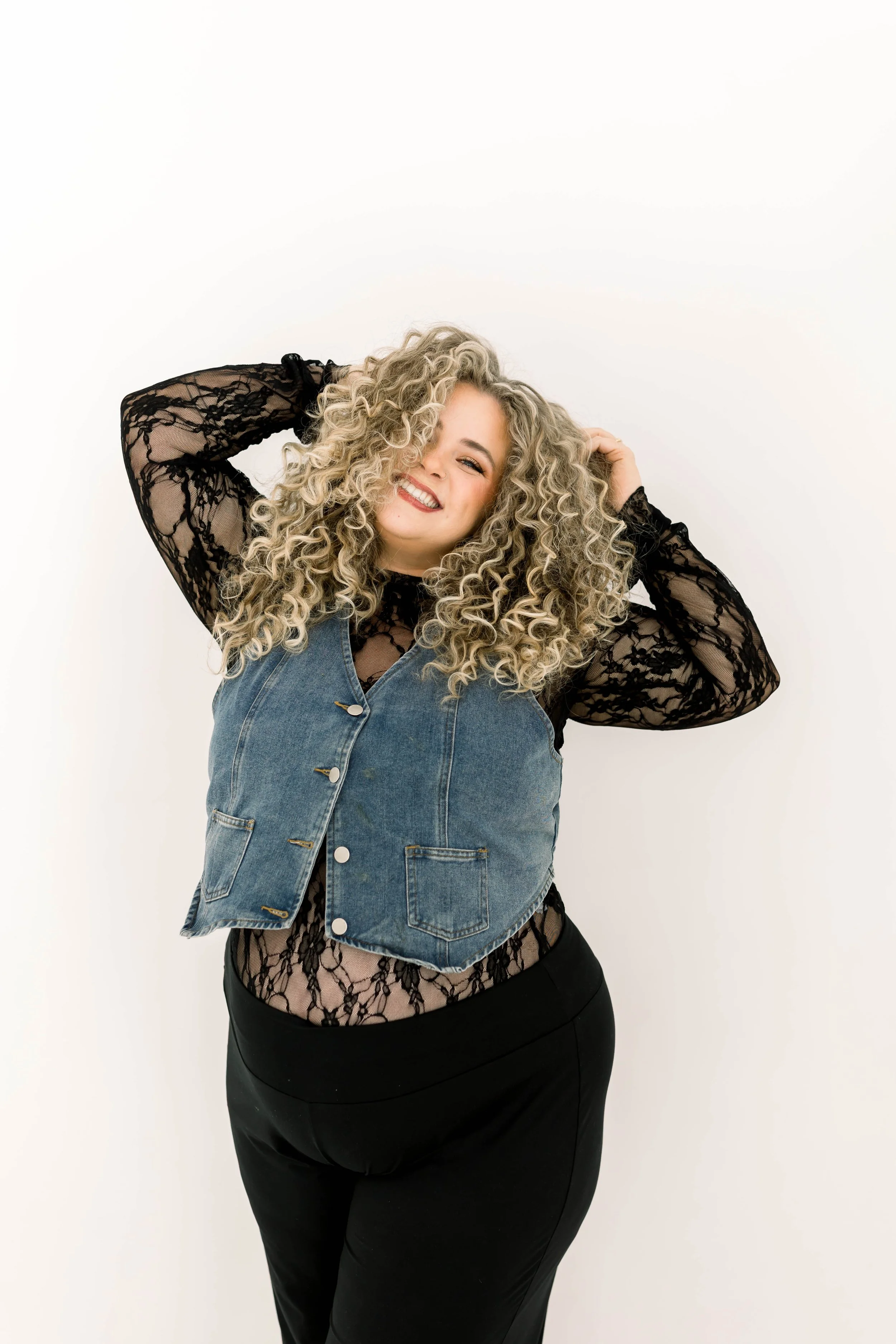Young woman with blonde wavy hair, smiling, wearing a black hat, black top, denim jacket, and necklace, against a pink background.