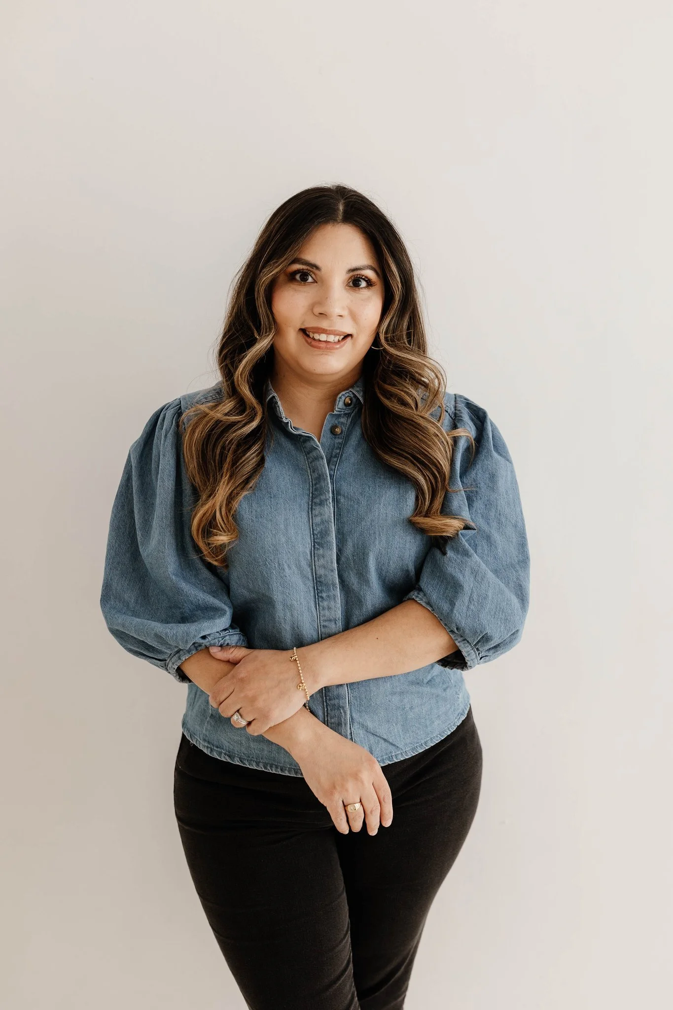 Portrait of a woman with long dark wavy hair, smiling, wearing a black top with sheer sleeves, against a pink background.