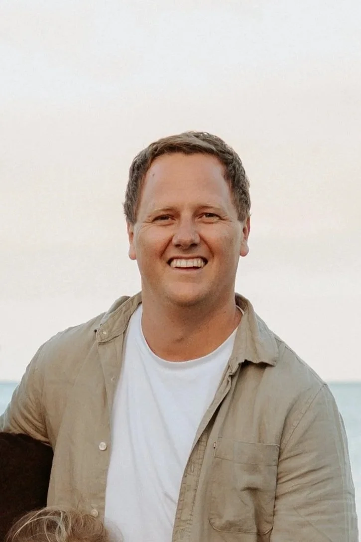A smiling man with short brown hair, wearing a beige button-up shirt over a white t-shirt, standing outdoors near the coast with an ocean in the background.