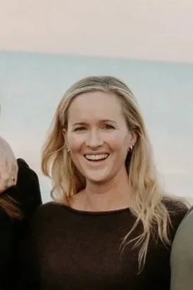 A woman with blonde hair smiling outdoors near the beach with water in the background.