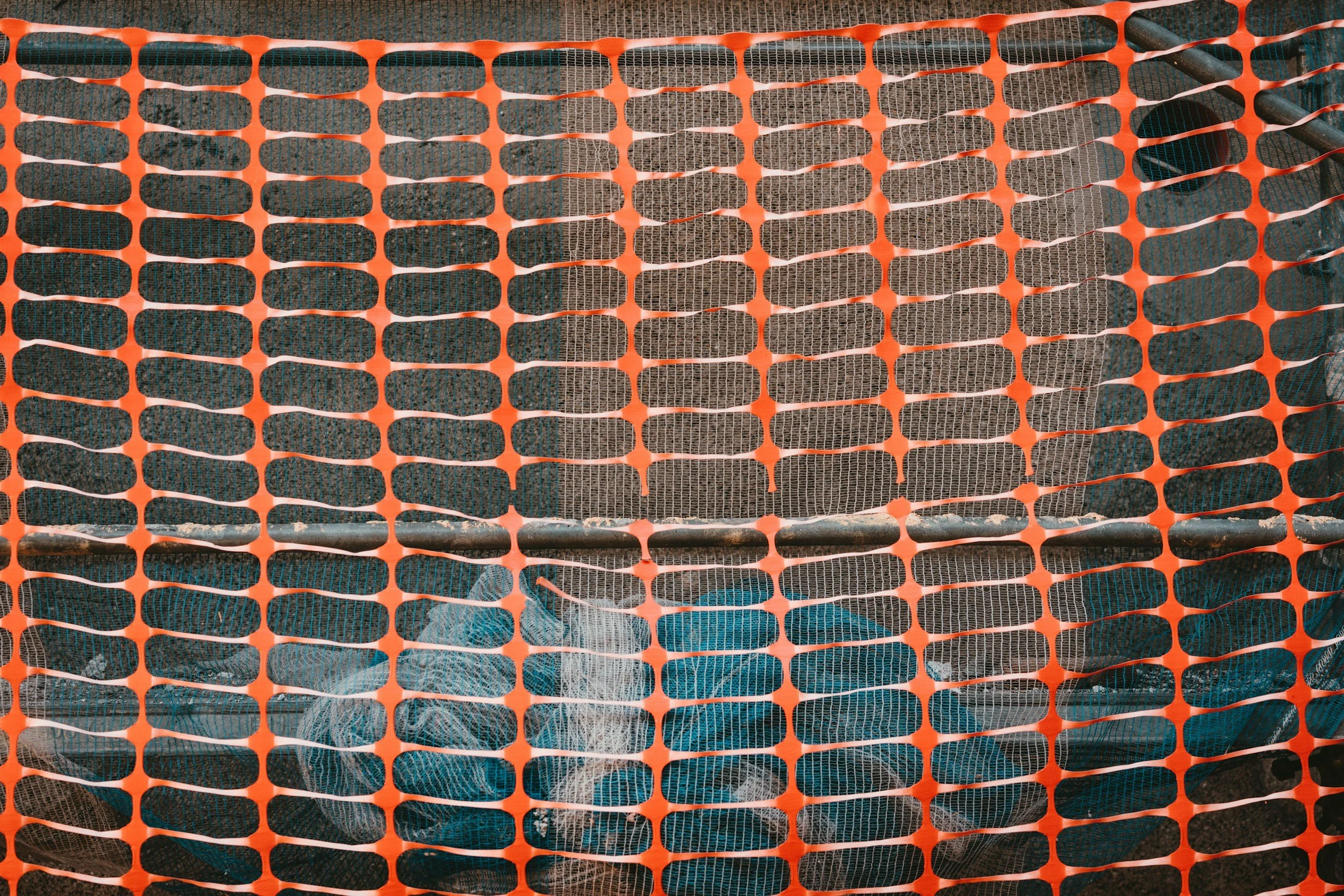 Tools and supplies behind orange construction safety netting, including mesh, tubes, and pipes.