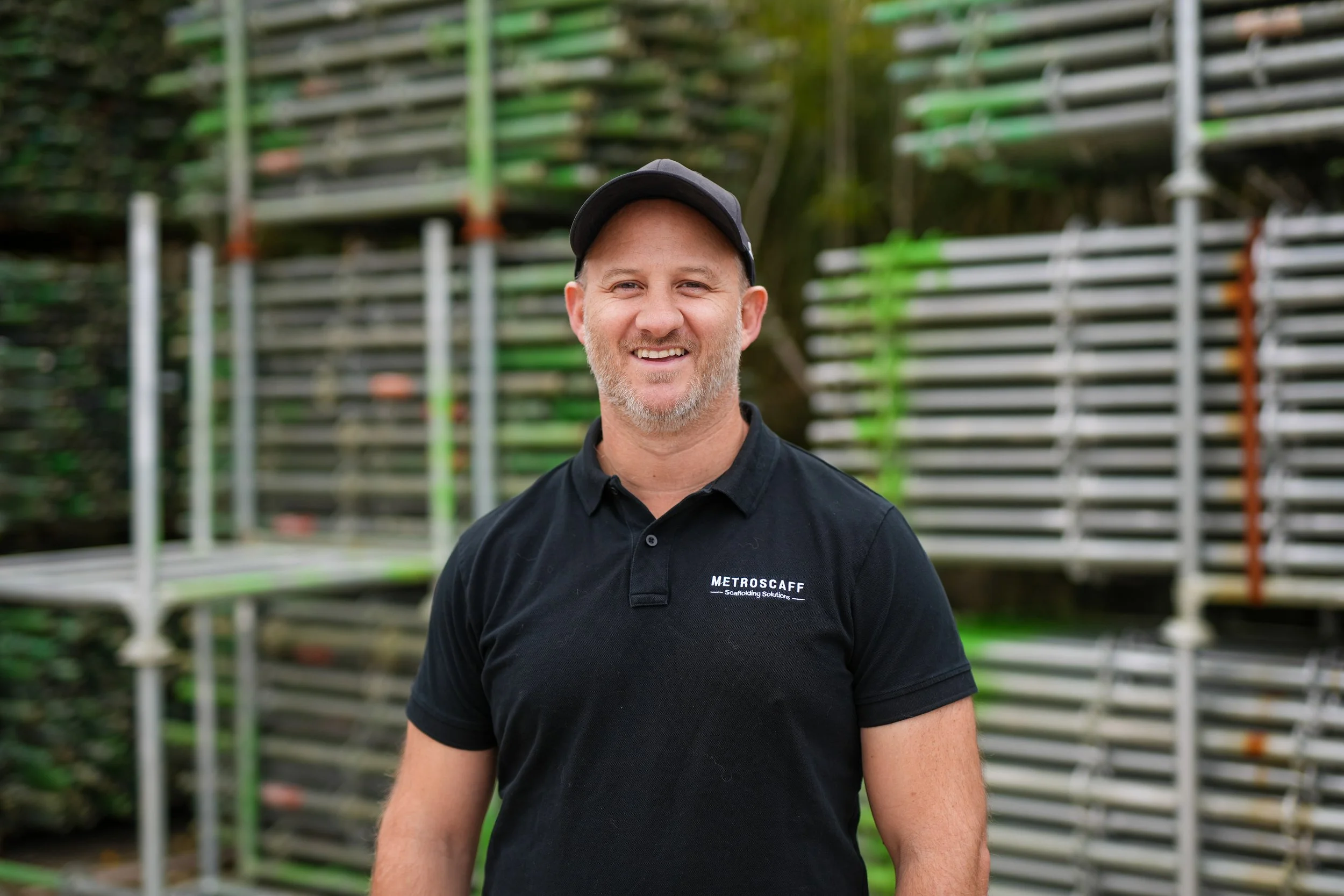 A man with a cap and beard, smiling, wearing a black polo shirt, standing outdoors near the ocean during sunset or sunrise.