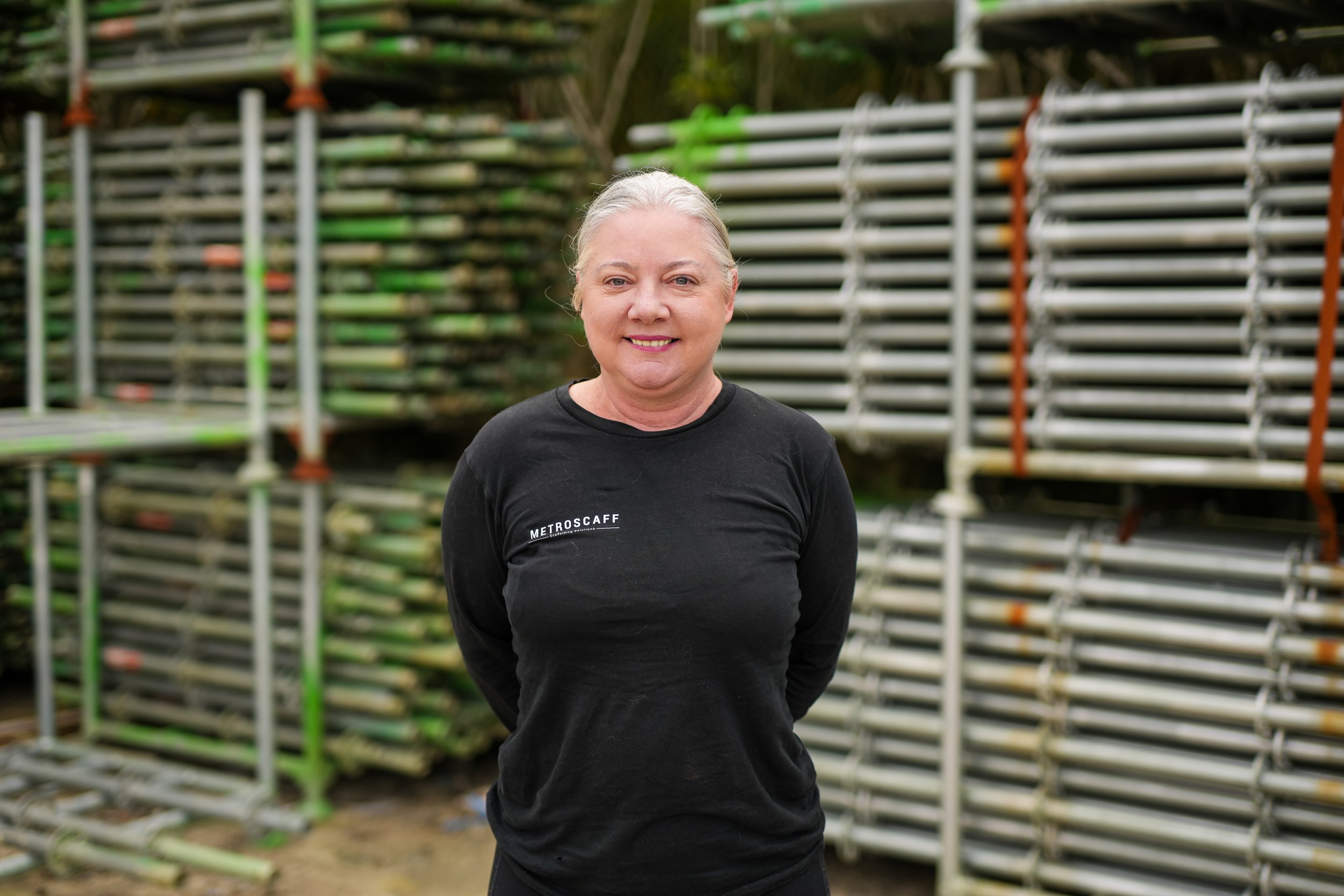 A smiling woman with blonde hair wearing a black top and a delicate necklace in a well-lit room with green plants and white shelves in the background.