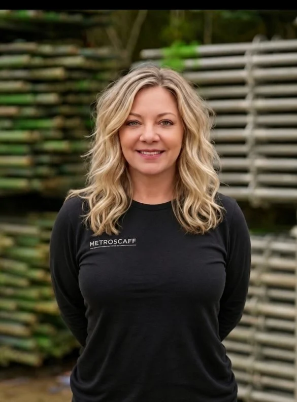 A smiling woman with blonde hair wearing a black top and a delicate necklace in a well-lit room with green plants and white shelves in the background.