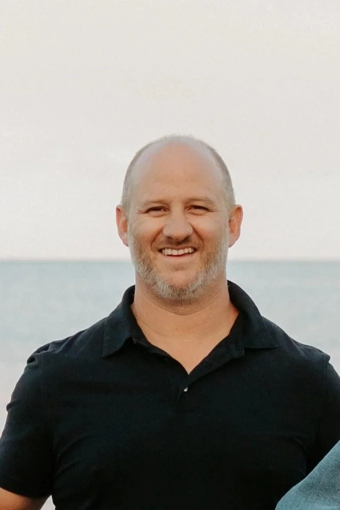 A man with a bald head and beard, smiling, wearing a black polo shirt, standing outdoors near the ocean during sunset or sunrise.