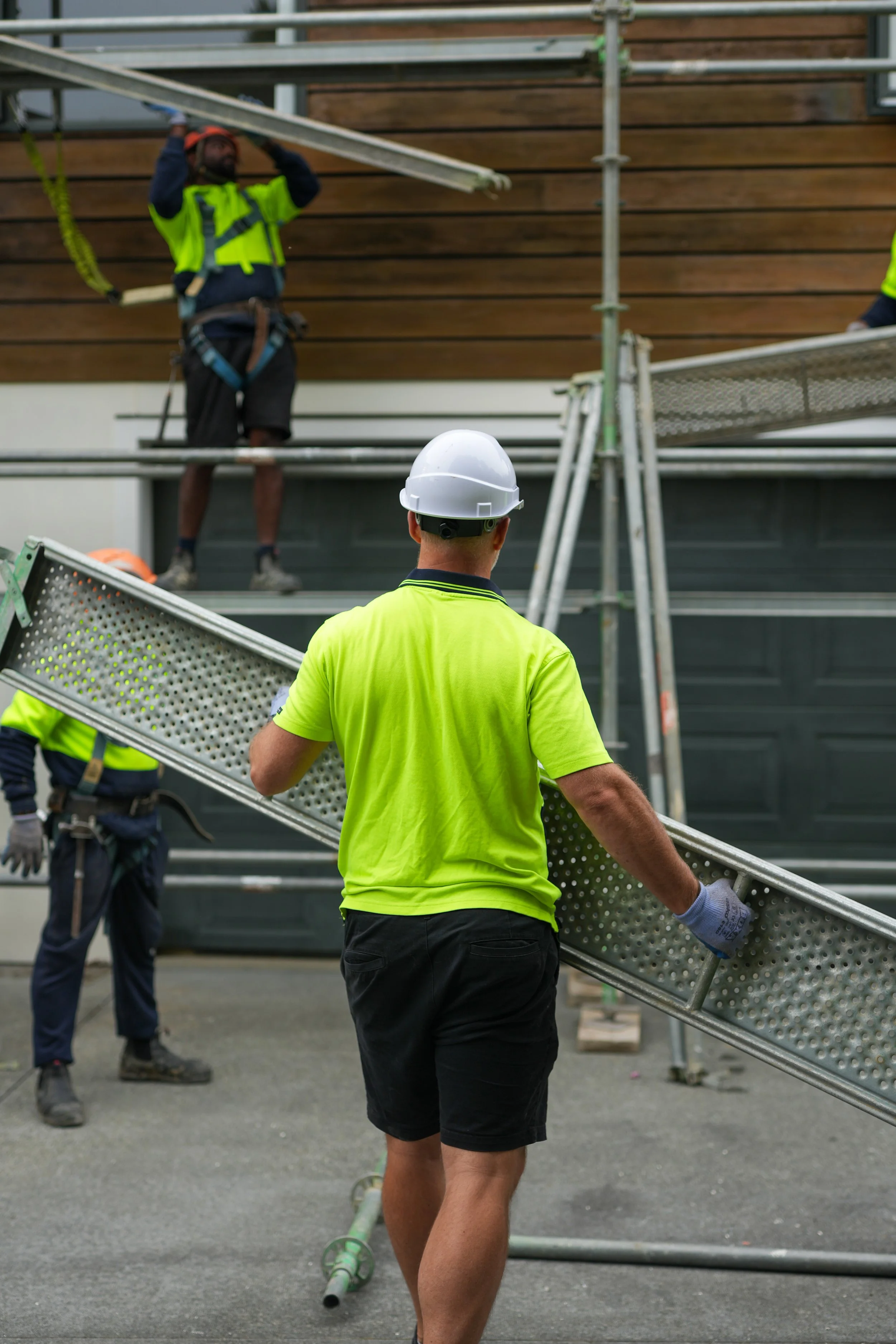 Scaffolders assembling scaffolding outside a building, with one worker in a white hard hat and bright yellow shirt guiding the kick board