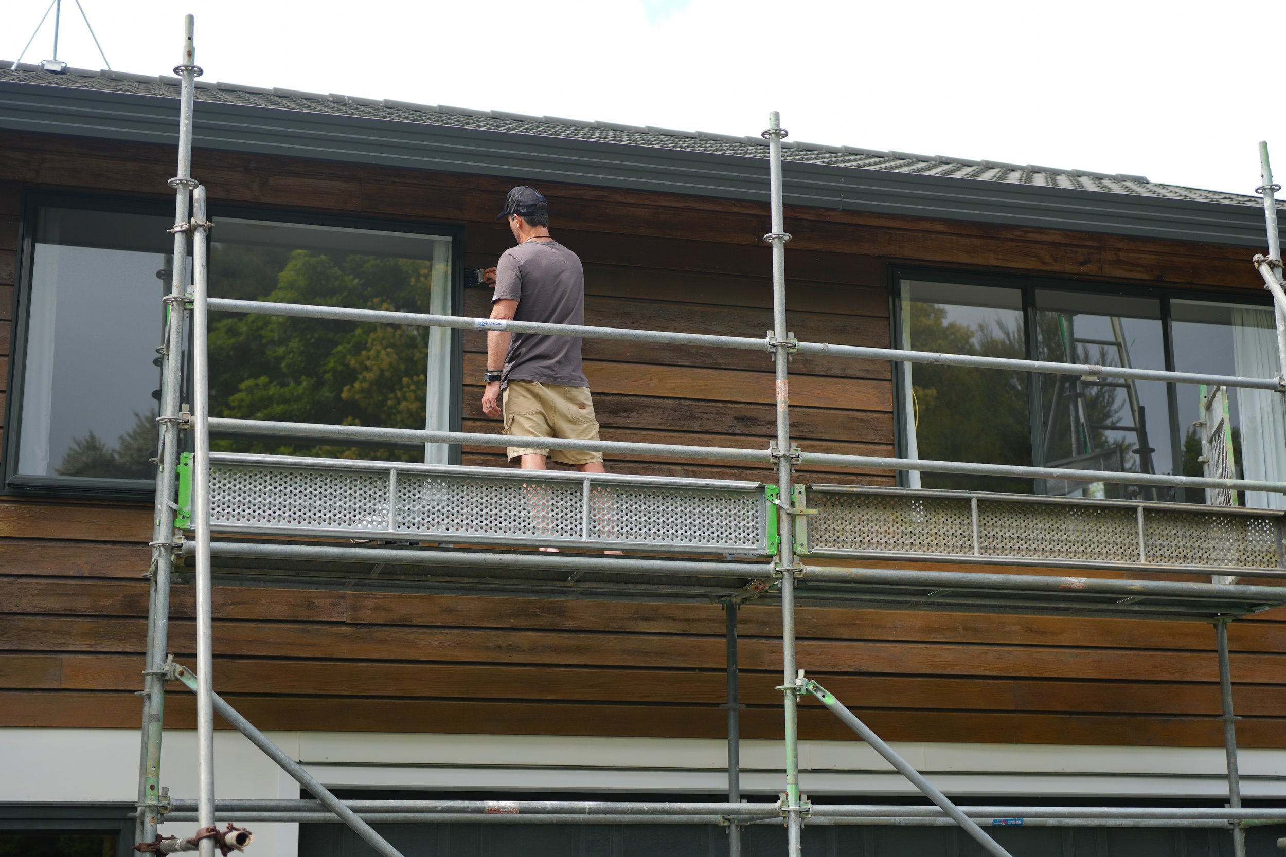 Man standing on scaffolding structure on the perimeter of a residential house painting the weatherboards
