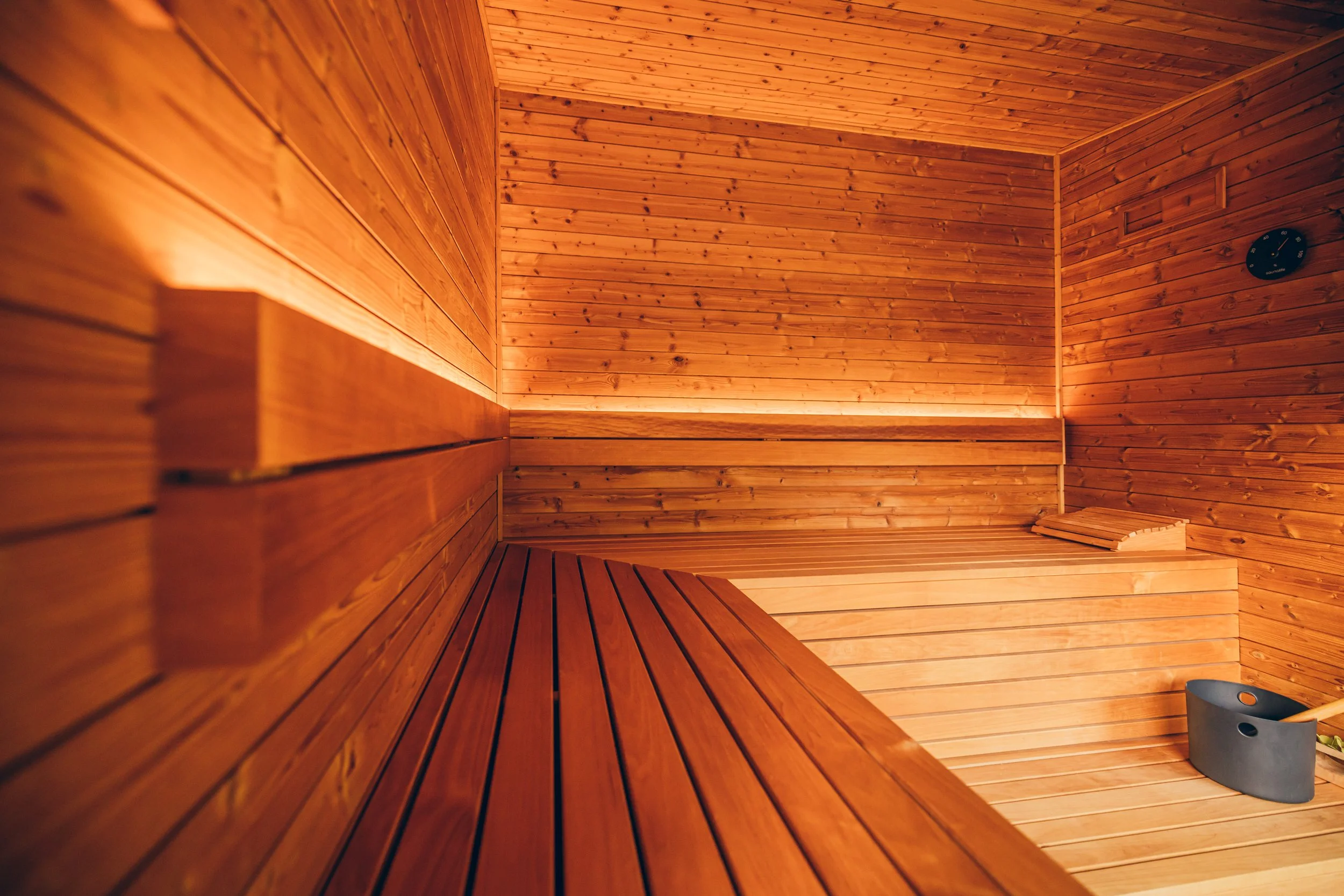 An empty wooden sauna with horizontal wooden planks on the walls and ceiling, a bench along the walls, a small clock on the wall, and a bucket on the floor.