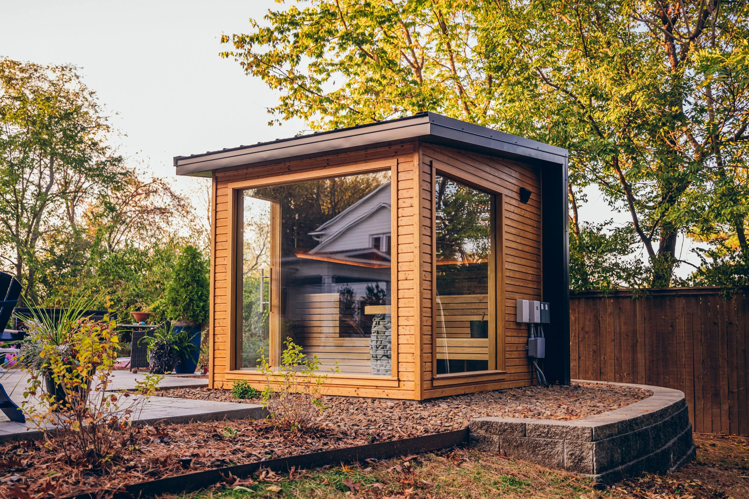 Chillfire Sauna Company small modern wooden garden sauna with large glass door and windows, surrounded by landscaped backyard with trees and wooden fence, designed for home wellness and relaxation.

