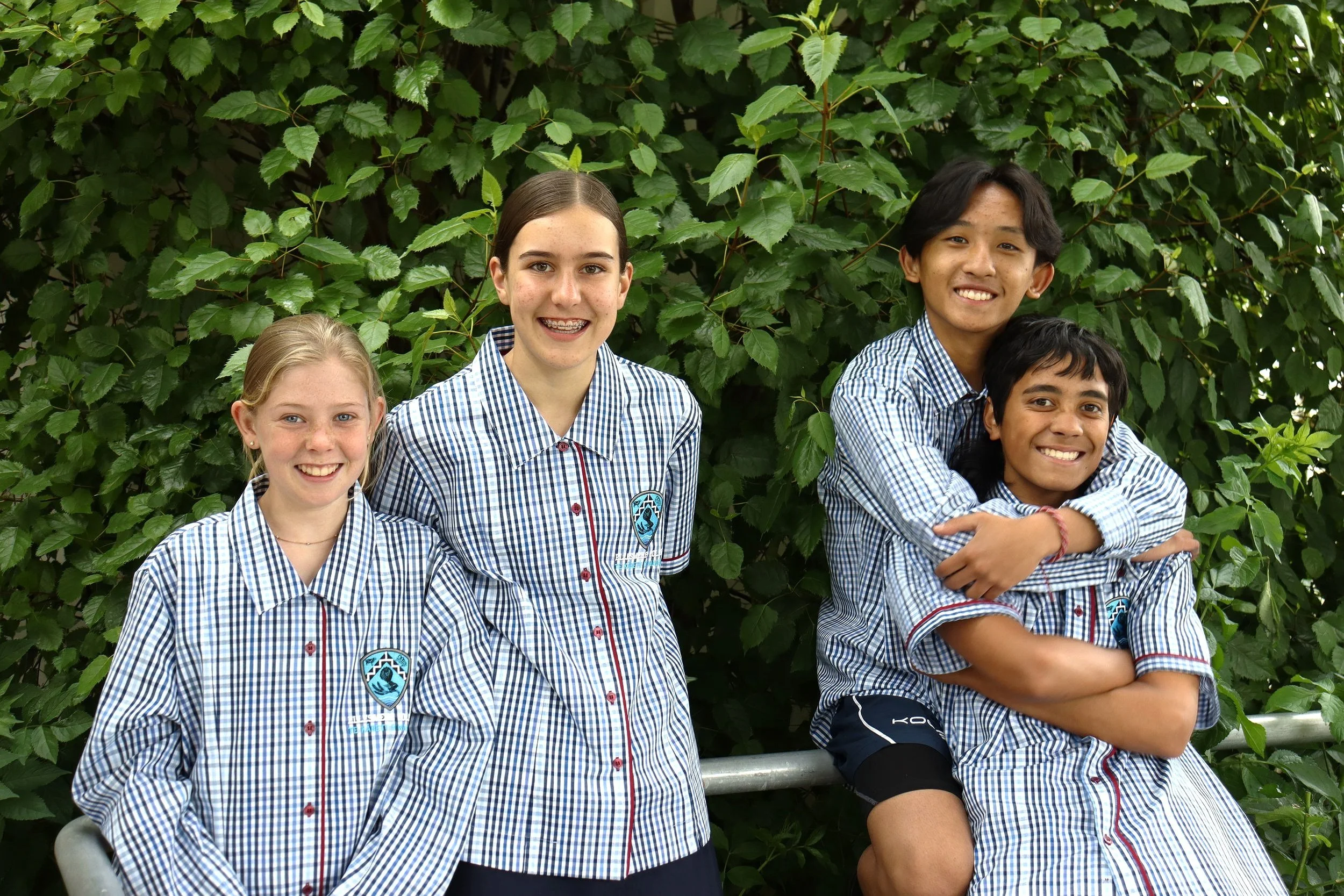 Four young students in school uniforms standing and sitting together outdoors in front of green foliage, smiling.