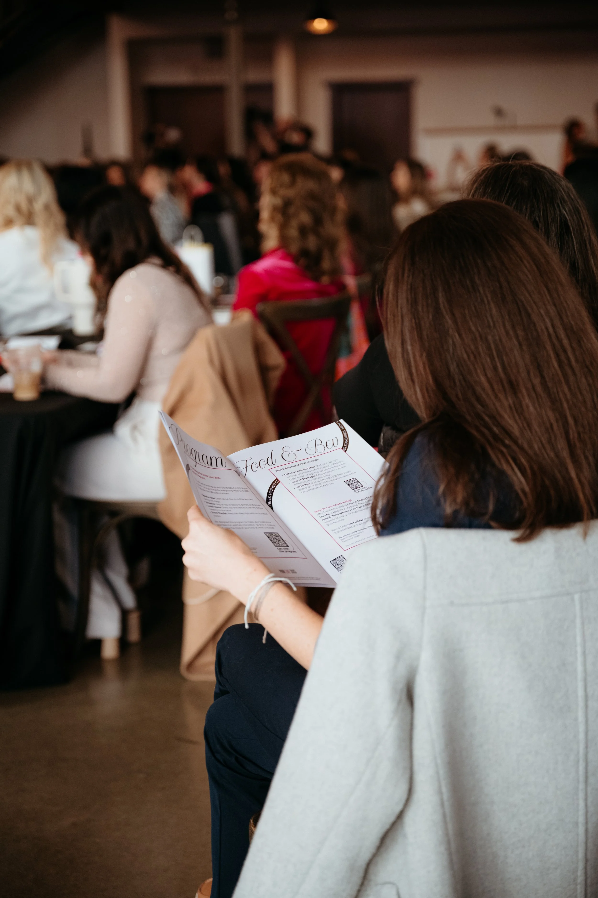 A woman with brown hair wearing a gray blazer is reading a food and beverage menu at a crowded event or conference with many people seated at tables.
