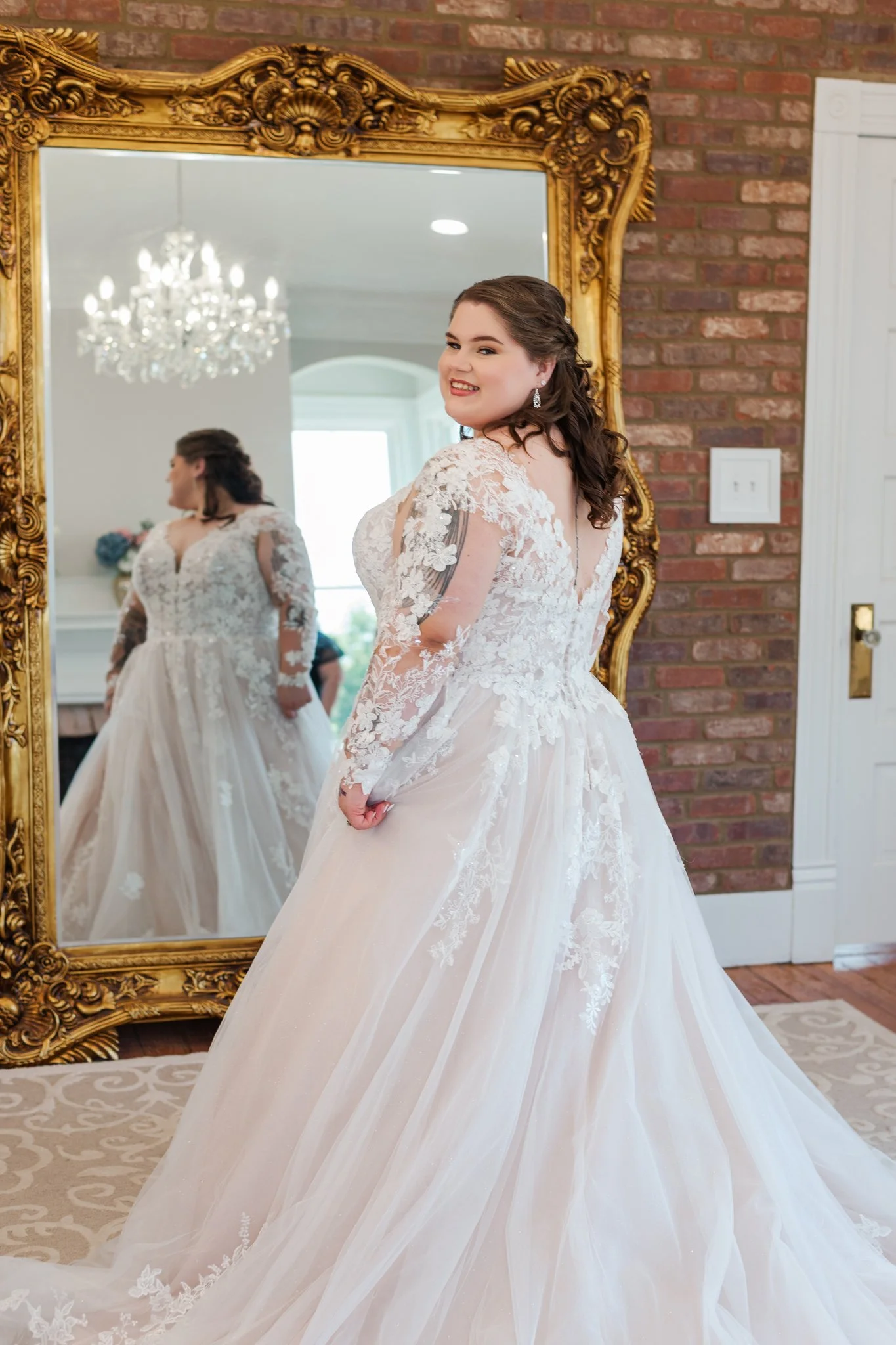 A woman in a wedding dress smiling at her reflection in a large ornate gold mirror in a room with a brick wall and a chandelier.