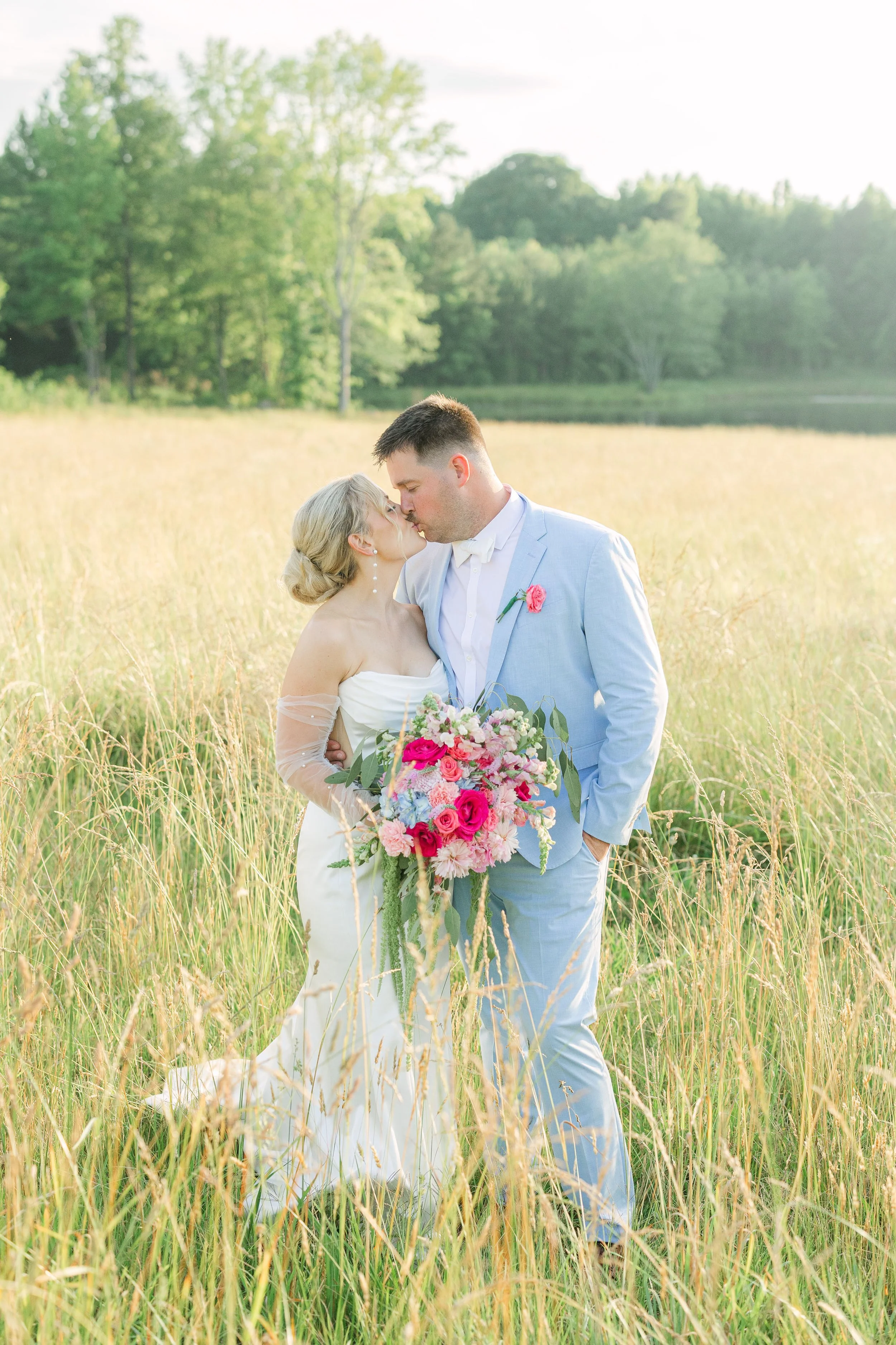 A bride and groom sharing a kiss in a field of tall yellow grass, holding a bouquet of pink and purple flowers, with a backdrop of trees and a pond.