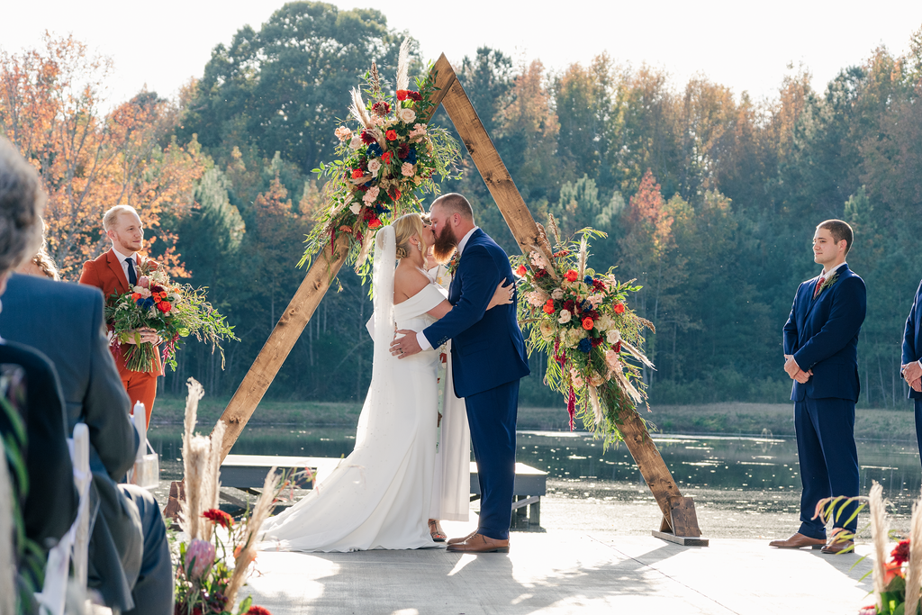 Couple kissing during outdoor wedding ceremony on a wooden platform beside a lake, with friends or family seated nearby, decorated with a large floral arch.