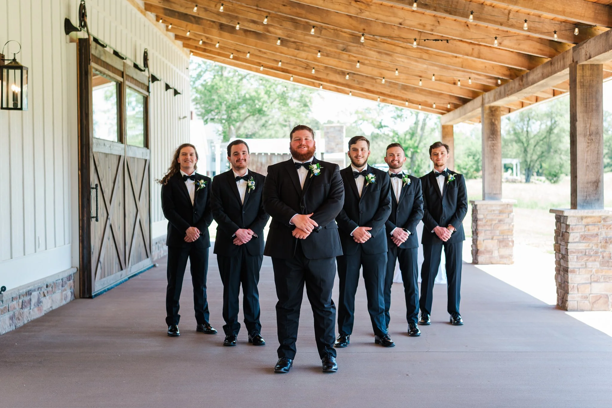 A group of six men in black tuxedos, standing on a covered outdoor porch with string lights and rustic wood and brick accents, smiling at the camera.