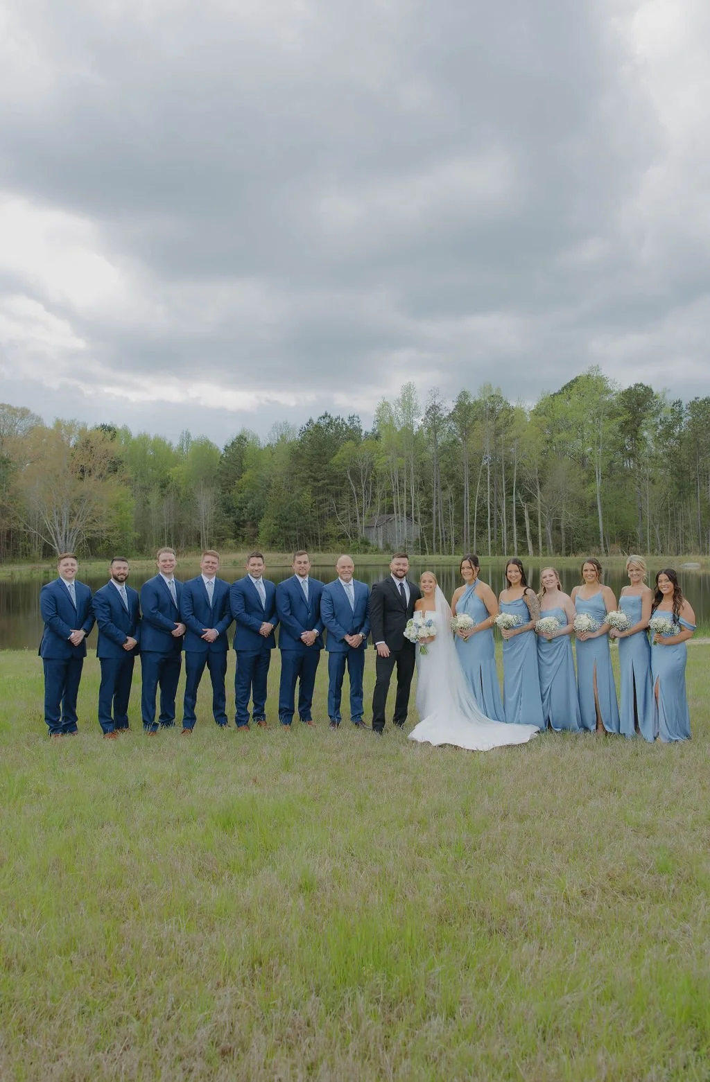 A wedding party standing outdoors by a lake with cloudy sky and green trees, comprised of groomsmen in blue suits, bridesmaids in light blue dresses, a bride in white gown, and a groom in black suit.