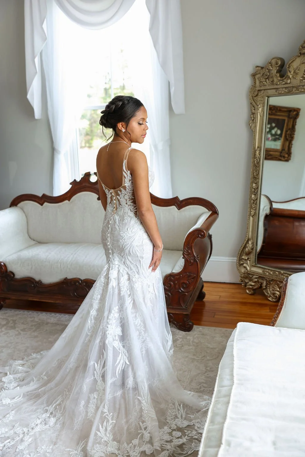 A bride in a white lace wedding dress standing in a vintage room with a large mirror and white curtains.