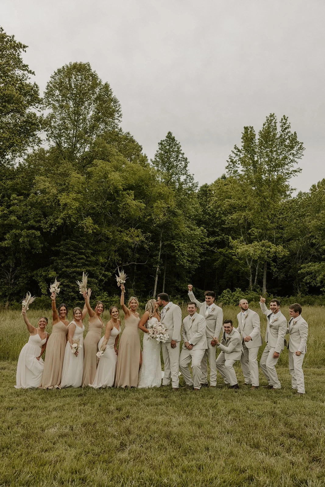 A group of wedding party members, with the bride and groom in the center, celebrating outdoors on grass with trees and an overcast sky in the background.