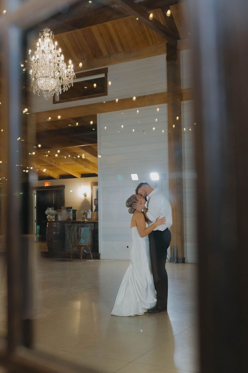 A bride and groom sharing a kiss during their wedding reception, seen through a window with warm lighting and elegant chandeliers reflected on the glass.