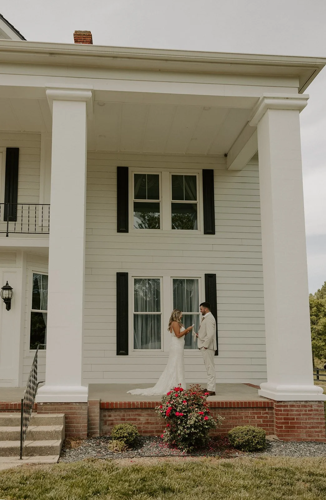 A couple dressed in wedding attire standing on the porch of a white house, with the bride reading from a paper or card to the groom.