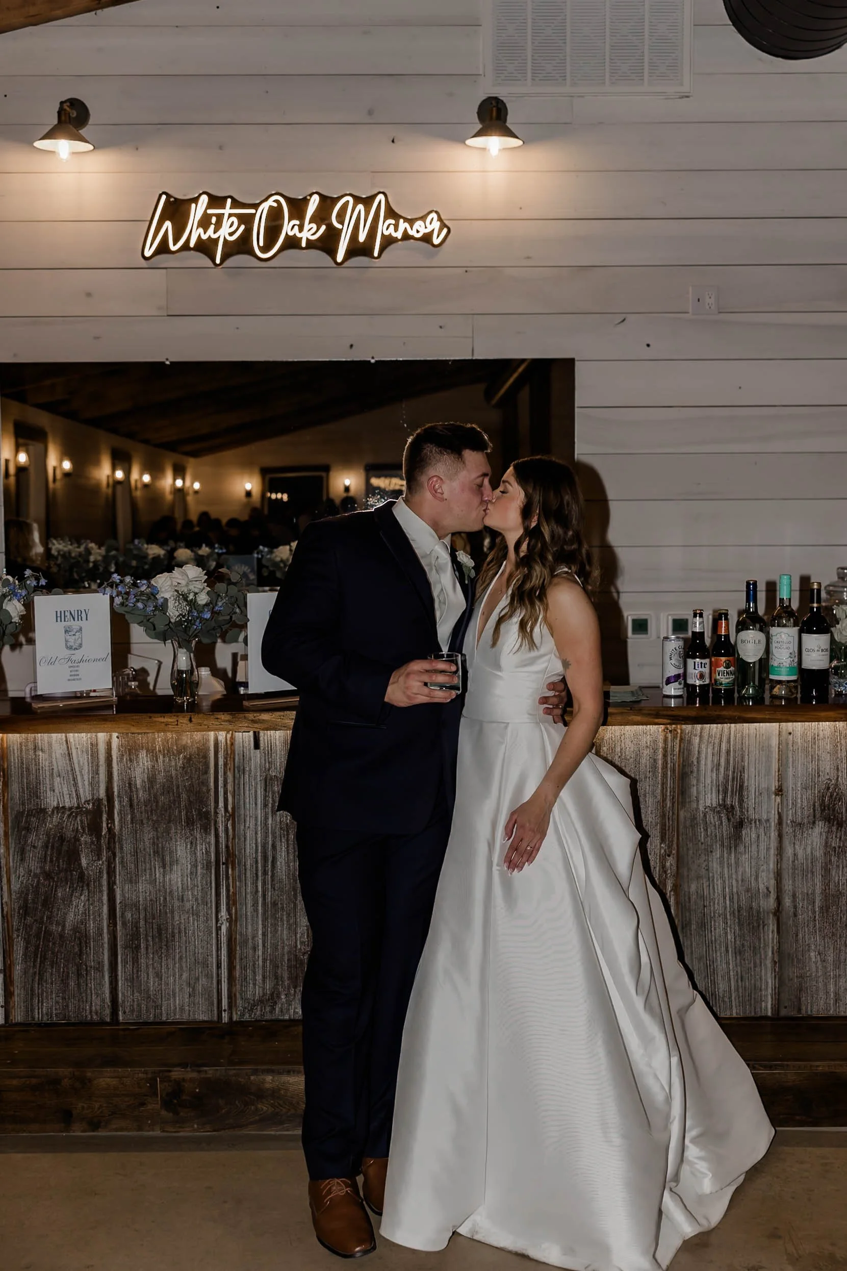 A newlywed couple sharing a kiss at their wedding reception in front of a bar, with a neon sign reading 'White Oak Manor' above them.