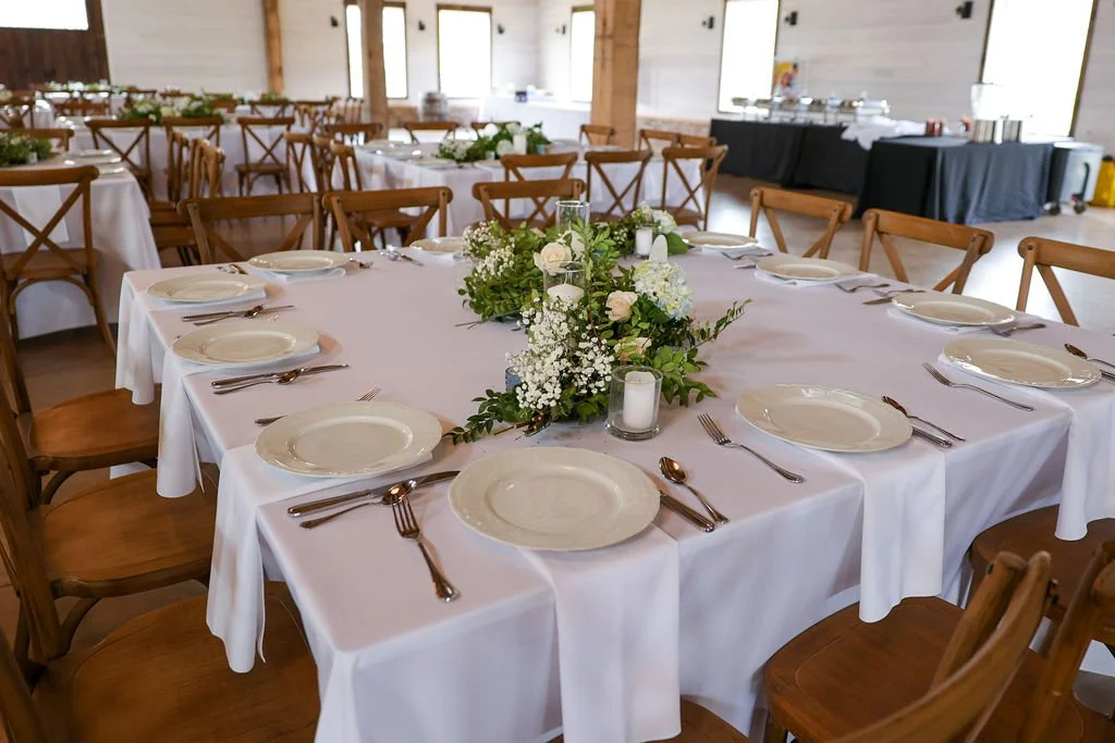 Wedding reception hall with round tables covered in white tablecloths, decorated with green and white floral centerpieces, and set with white plates, silverware, and candles.