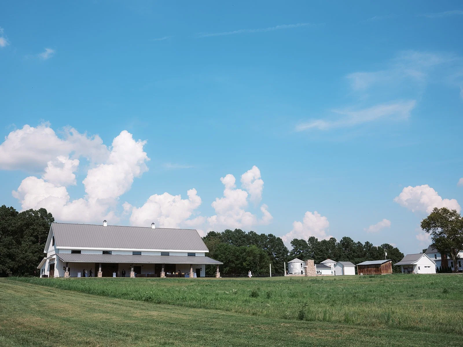 A rural landscape with a large barn, green fields, and several smaller white buildings against a bright blue sky with white clouds.