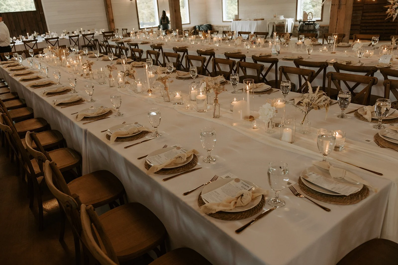 A long wedding reception table decorated with candles, flowers, glassware, and table settings in a rustic wooden venue.