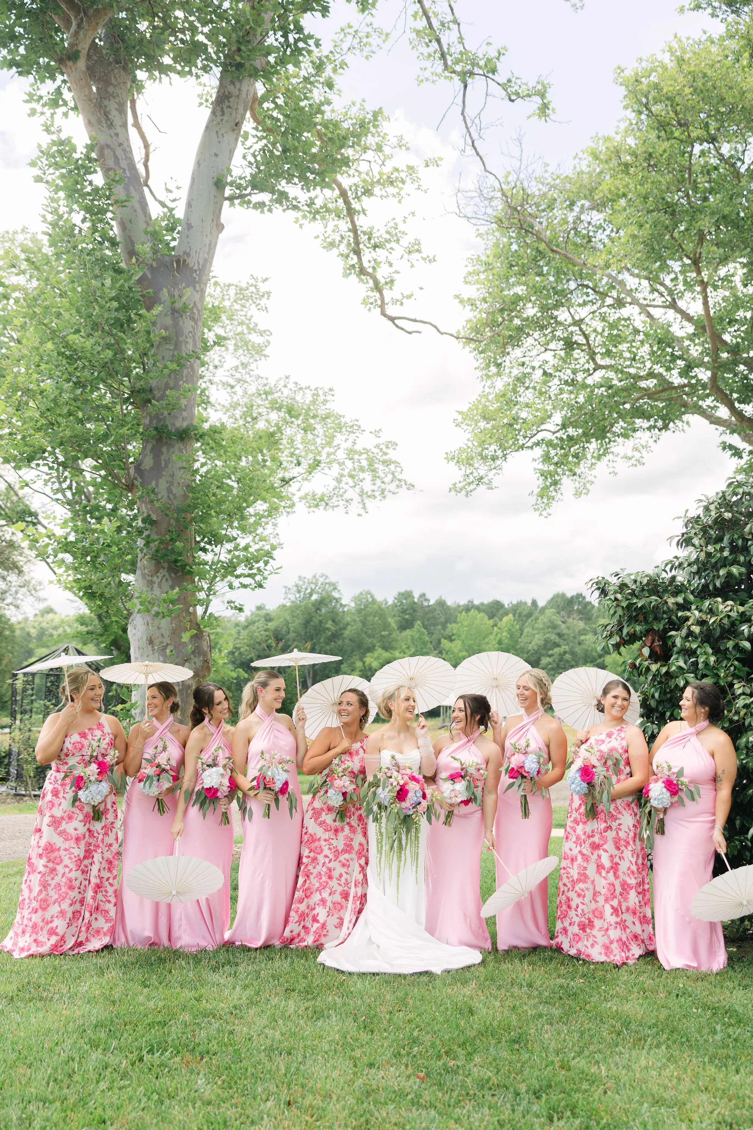 Bridesmaids and bride at an outdoor wedding, all holding pink and purple floral bouquets and white parasols, standing on grass under trees with a cloudy sky.