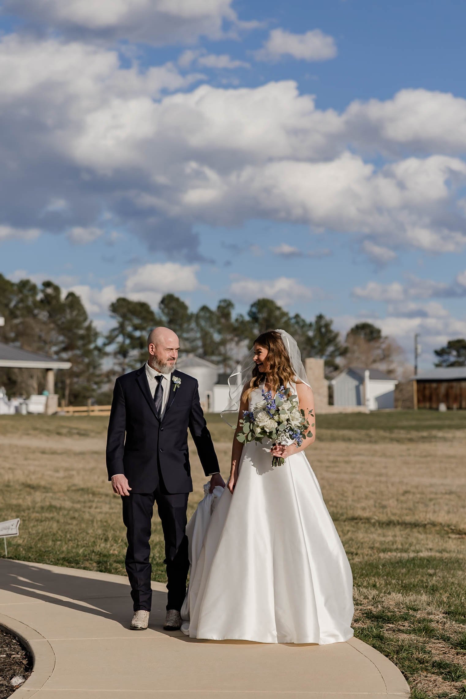 A bride in a white wedding dress holding a bouquet, walking with a man in a black suit on a sidewalk outdoors with cloudy sky and trees in the background.