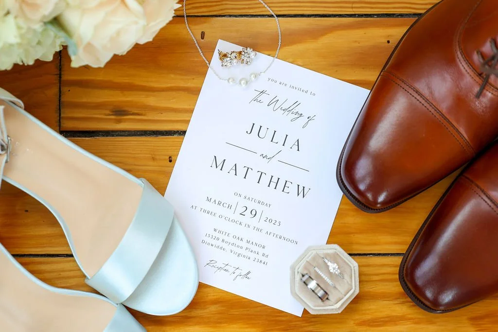 Wedding invitation surrounded by a pair of brown leather shoes, light blue sandal, wedding rings in a small hexagonal box, a pearl necklace, and white flowers on a wooden surface.