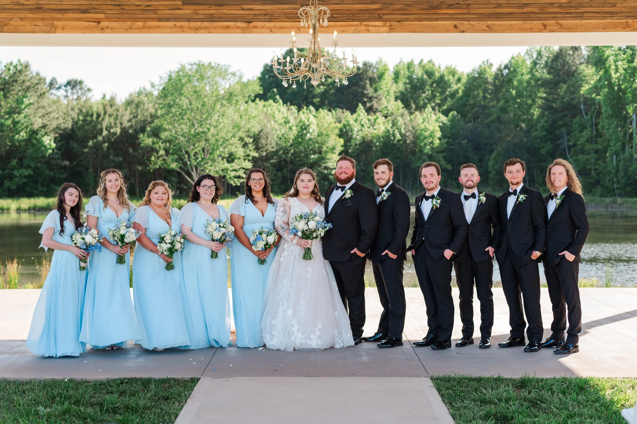 A wedding party of ten people standing outdoors near a lake and green trees, dressed in formal attire with the bride and groom in the center, the women in light blue dresses and the men in black tuxedos.
