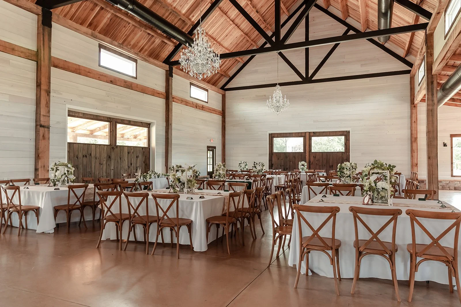 Interior of a rustic wedding or event hall with round tables covered in white tablecloths, decorated with floral centerpieces, elegant chandeliers hanging from a wooden ceiling, and large windows for natural light.
