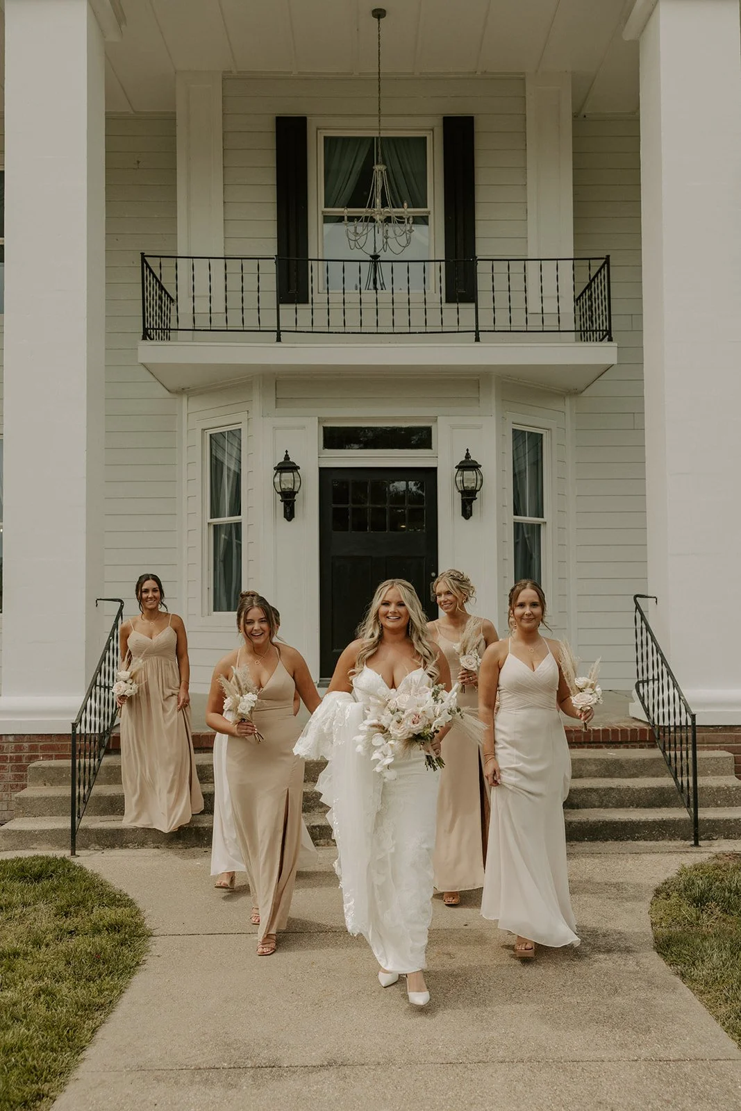 A bride and five bridesmaids walking down the steps of a large white house, with the bride in a white wedding gown holding a large bouquet and the bridesmaids in beige dresses carrying smaller bouquets.