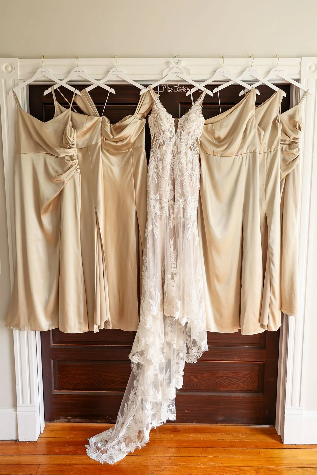 A group of six beige bridesmaid dresses and one white lace wedding gown hanging on white hangers on a dark wooden door.