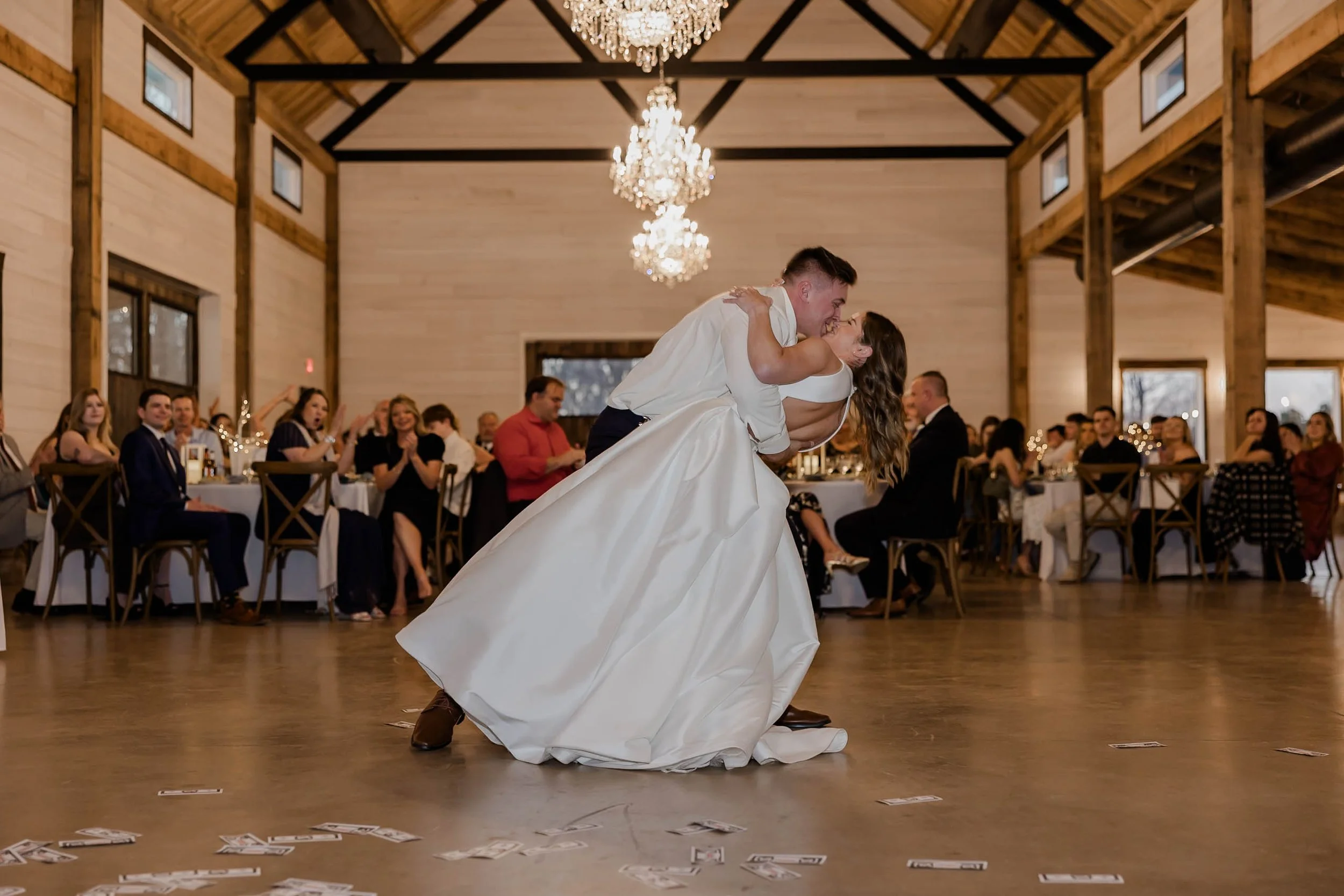 A bride and groom share a dance during their wedding reception. The groom is dipping the bride as they smile at each other. Guests sit at tables decorated with lights, watching the couple, with some clapping and smiling.