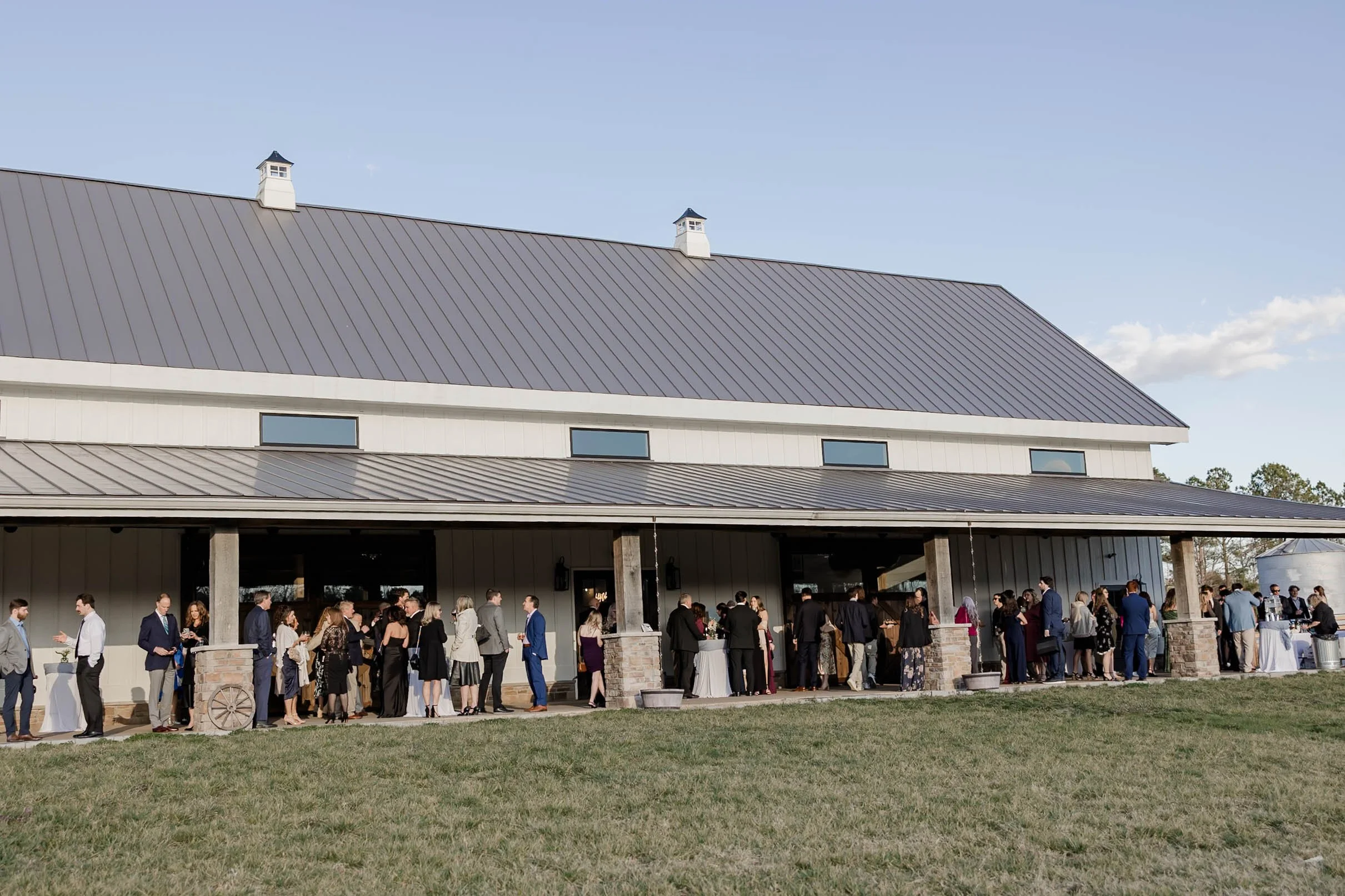 People at an outdoor event in front of a large beige building with a metal roof and small windows, under a clear blue sky.
