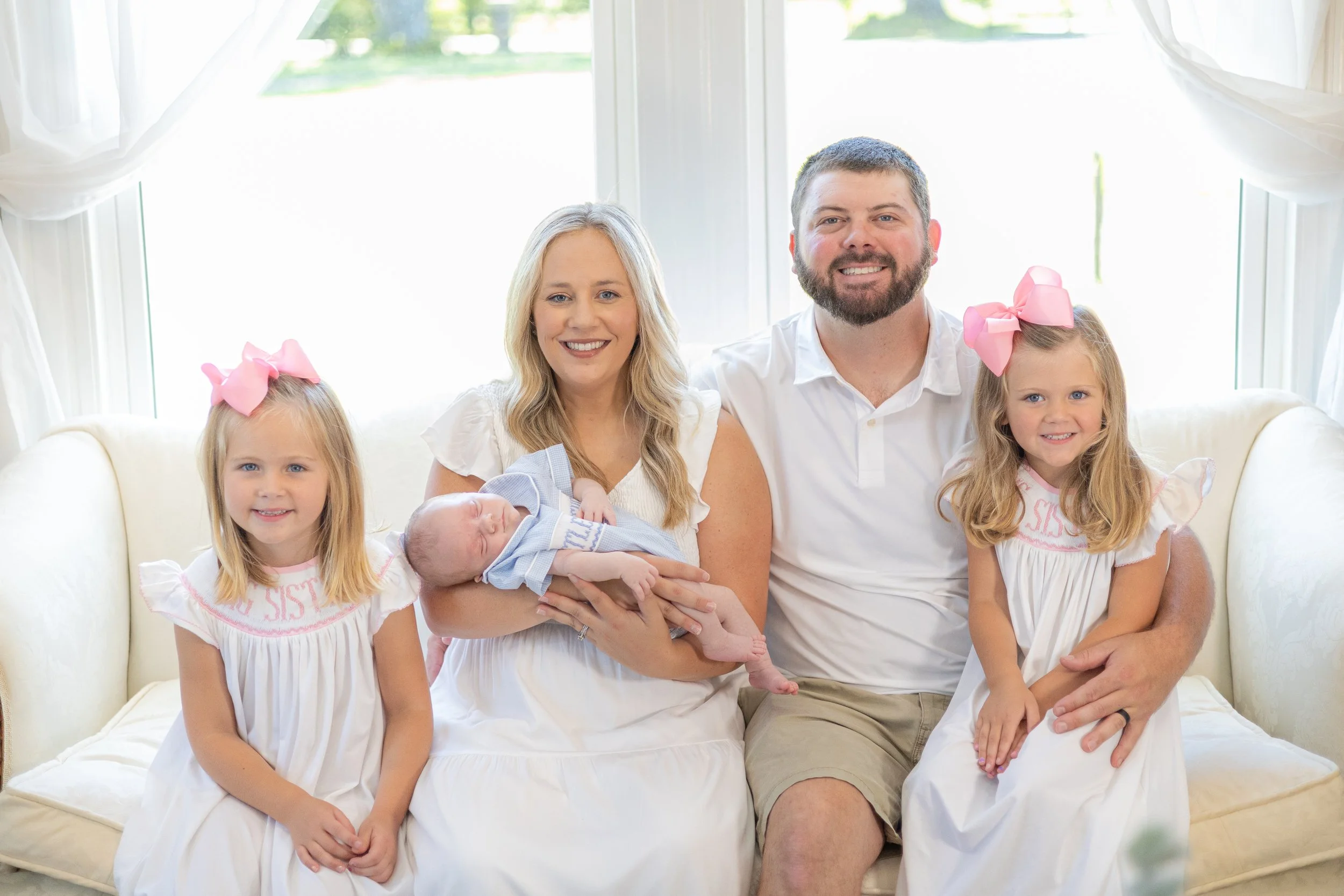 Family of five sitting together on a white couch in bright room, with two young girls, a woman holding a newborn, and a man, all smiling at camera.