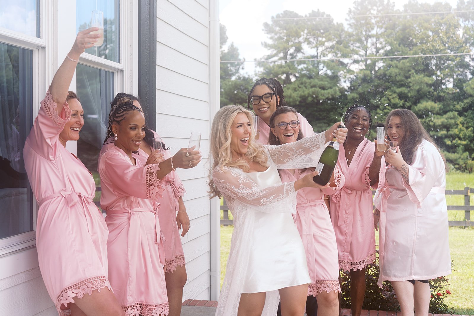 A bride and friends celebrating outdoors on a porch, with the bride holding a champagne bottle and glasses raised, all smiling and wearing pink robes.