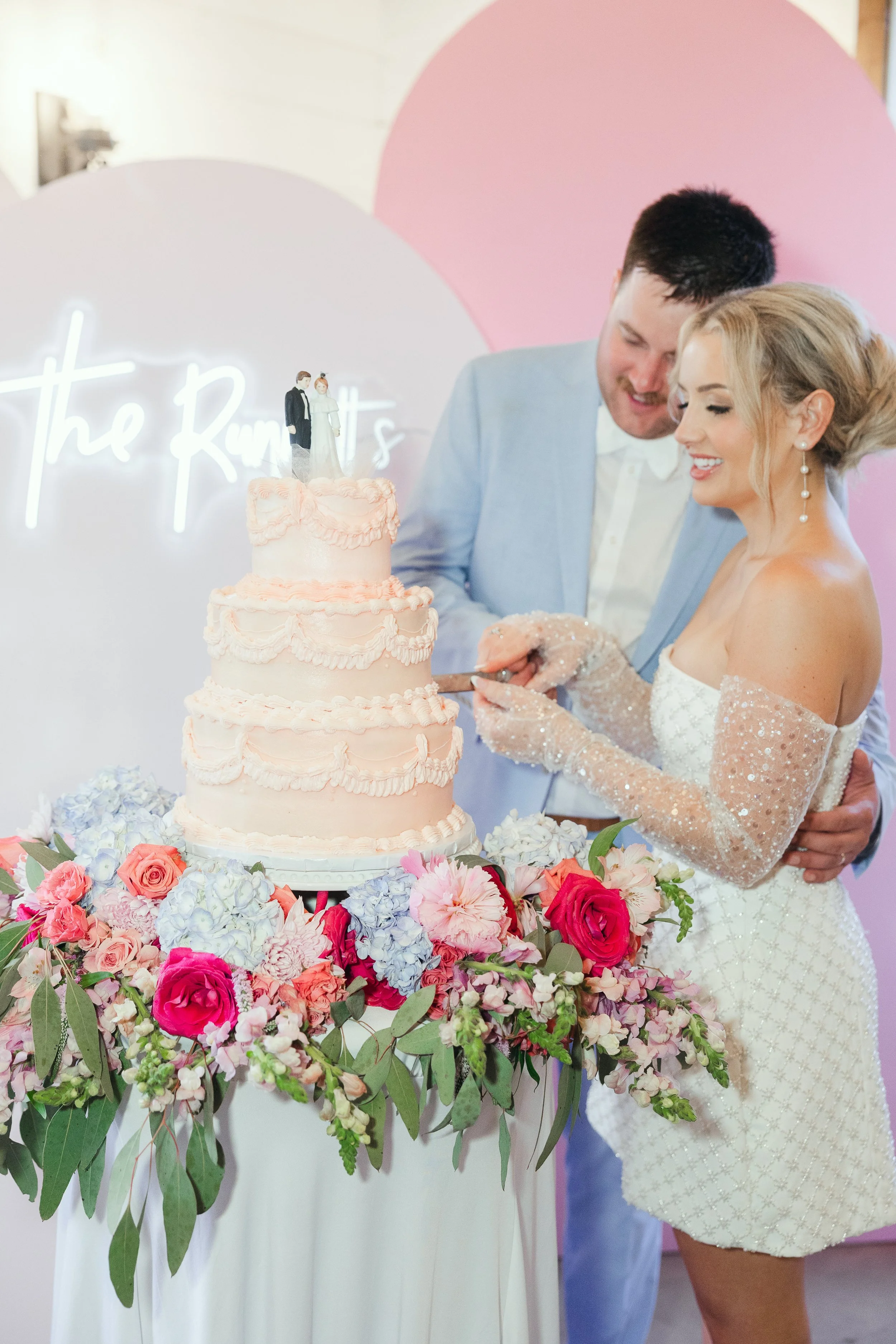 A bride and groom cutting their wedding cake together, standing next to a table decorated with pink and purple flowers.