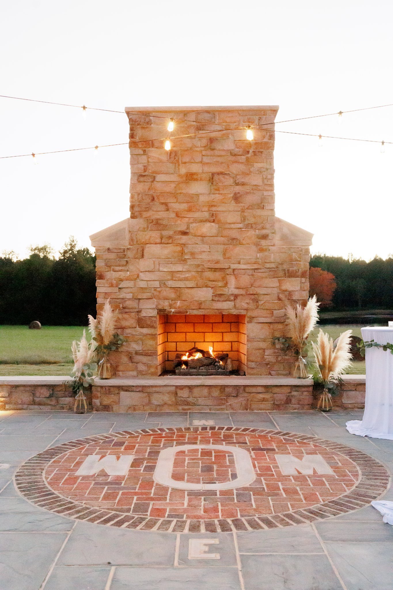 Outdoor brick fireplace decorated with pampas grass vases, with a fire burning and string lights overhead, set on a patio with a circular brick and stone design reading 'WOMEN'.