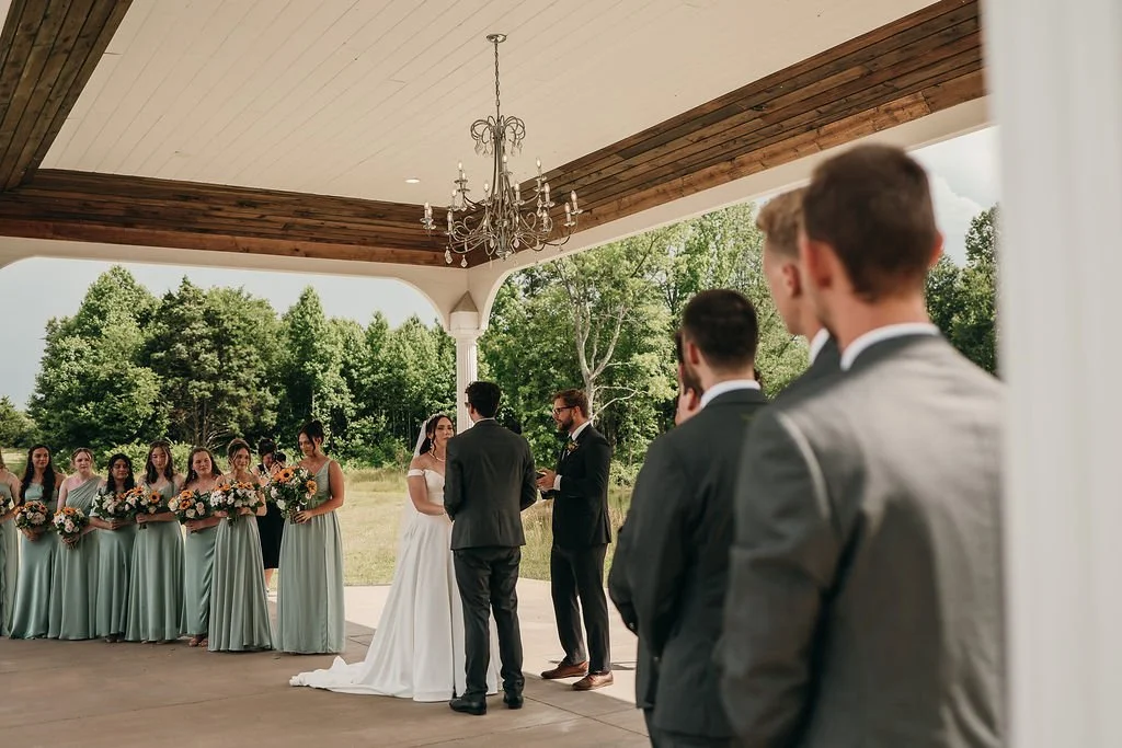 A wedding ceremony taking place outdoors under a pavilion, with a bride and groom standing in front of a officiant, surrounded by bridesmaids and groomsmen.
