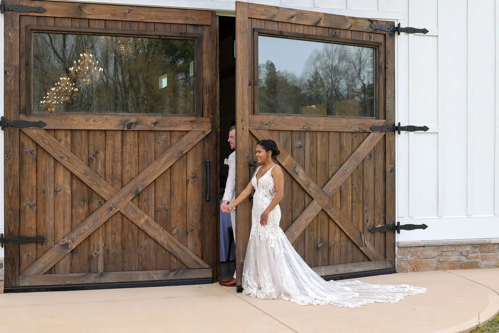 A bride in a white wedding gown is standing outside a large wooden barn door, holding hands with a groom who is partially hidden behind the door.