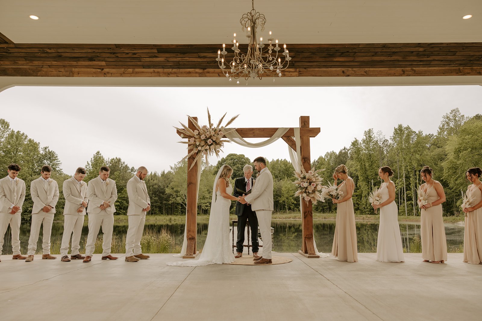 Wedding ceremony outdoors by a lake with a wooden arch, bride and groom facing each other holding hands, officiant standing behind them, six groomsmen on the left in white suits, six bridesmaids on the right in light-colored dresses, all holding bouquets, surrounded by trees, under a chandelier and wood ceiling, during daytime.