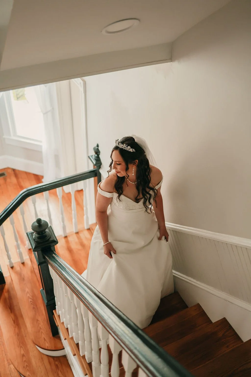 A bride in a white wedding gown standing on a staircase with wooden steps and a black and white railing, looking down and smiling, wearing a tiara, earrings, and a necklace, with a veil and dark curly hair.