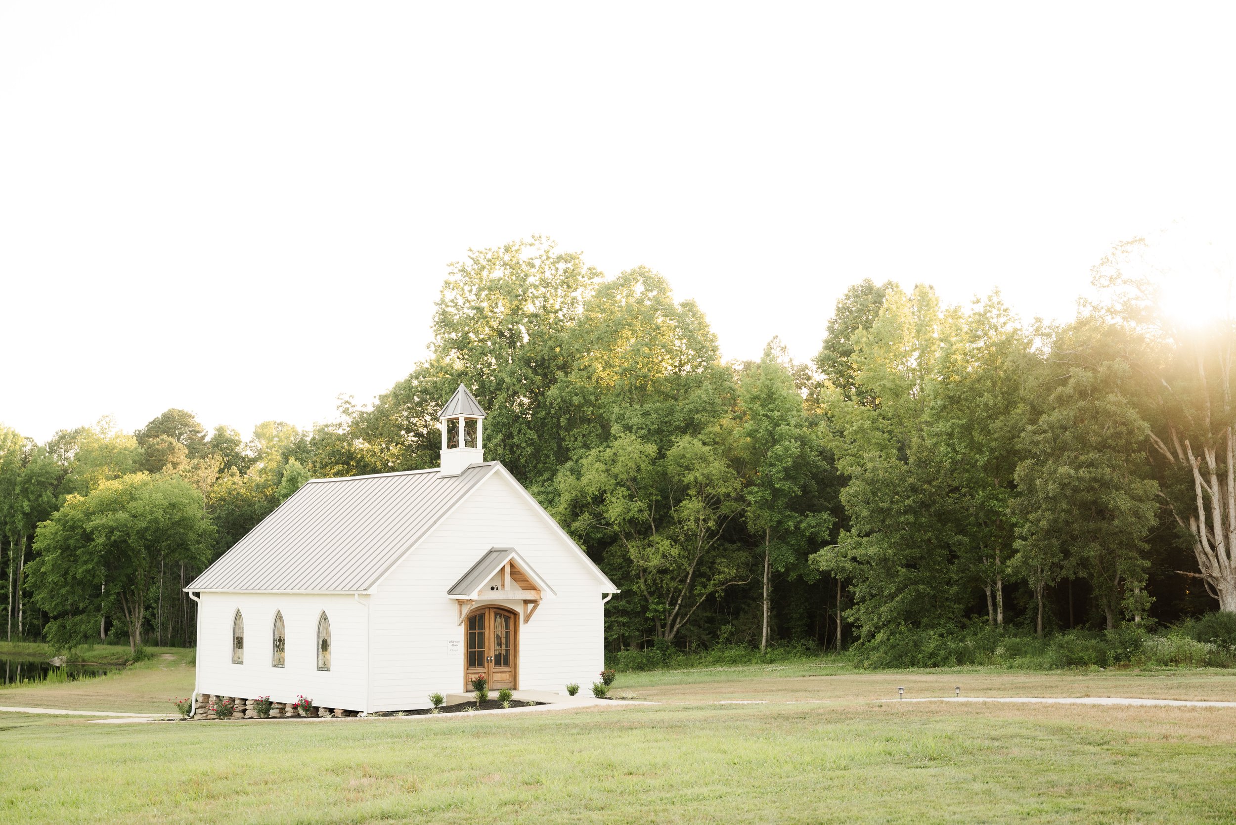 A small white chapel with a wooden door, set in a grassy field with trees in the background and sunlight shining from the right.
