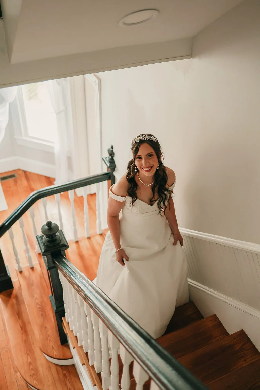 A bride smiling on a staircase, wearing a white wedding dress, tiara, pearl necklace, and earrings.